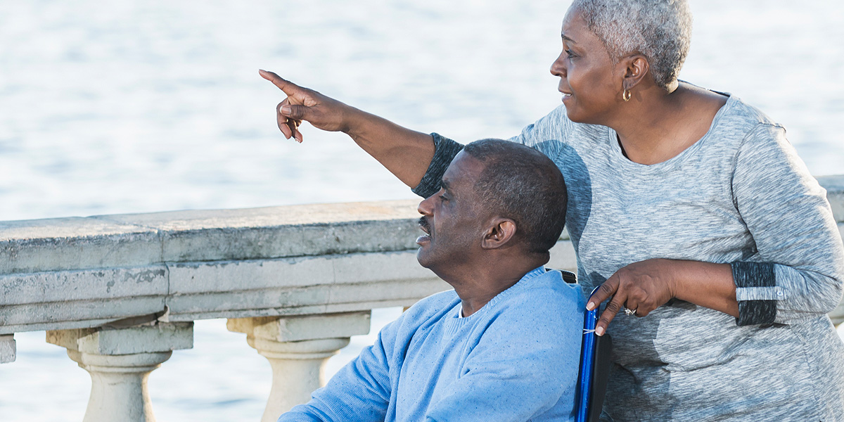 Image of elderly woman pointing to the sea and standing behind an elderly man in a wheelchair  Text: Busy lives. Places to go. People we care for.